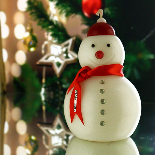 Festive snowman-shaped candle with a red scarf and a Christmas tree in the background.